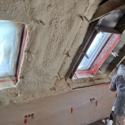 P.H.U Emilka Emilia Wołczyńska - Aplikacja izolacji pianką na poddaszu, widoczne okna dachowe i folia ochronna na ścianach, pracownik w kombinezonie ochronnym w trakcie natrysku.