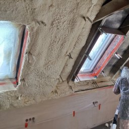 P.H.U Emilka Emilia Wołczyńska - Poddasze ocieplone pianką, widoczne okna dachowe i folia ochronna na ścianie, fragment drabiny i osoba w kombinezonie ochronnym.