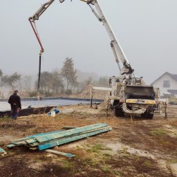 P.H.U Emilka Emilia Wołczyńska - Wylewanie betonu do przygotowanego wykopu pod fundamenty domu jednorodzinnego, widoczny dźwig z pompą do betonu, dwaj pracownicy kontrolują proces, w tle budynek w budowie i przenośna toaleta.