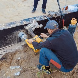 P.H.U Emilka Emilia Wołczyńska - Pracownik w trakcie wykonywania otworu w elemencie konstrukcyjnym przy użyciu wiertnicy diamentowej, widoczny fragment fundamentu i drugi pracownik w tle.