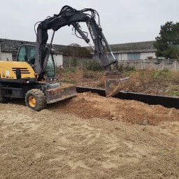 P.H.U Emilka Emilia Wołczyńska - Żółto-czarna koparka Mecalac wyrównuje teren pod budowę, widoczne przygotowane czarne szalunki, w tle zabudowania gospodarcze.