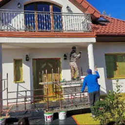 Dwaj pracownicy na rusztowaniu nanoszą tynk na elewację domu jednorodzinnego z zabezpieczonymi oknami i drzwiami, widoczna kolumna i balkon z metalową balustradą.