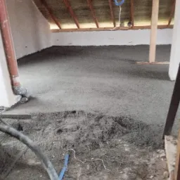 Świeżo wylana, mokra, szara masa na podłodze poddasza, widoczny wąż do pompowania mieszanki i drewniany szkielet dachu.