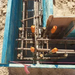 Zbrojenie fundamentu w szalunku z niebieskiego drewna, zabezpieczone rury w pomarańczowych osłonach, widoczny fragment buta pracownika.