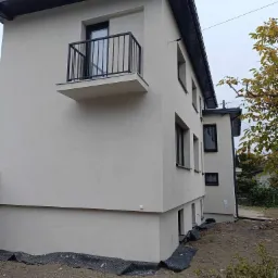 Nowoczesny, dwukondygnacyjny dom z jasną elewacją i czarnymi ramami okiennymi oraz balkonem z metalową balustradą, widok z zewnątrz na etapie zagospodarowania terenu.