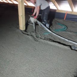 P.H.U Emilka Emilia Wołczyńska - Pracownik w szarych ubraniach i czerwonych rękawicach rozprowadza masę na podłodze poddasza za pomocą specjalistycznego urządzenia, widoczne drewniane elementy konstrukcji dachu.