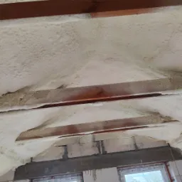 Poddasze ocieplone pianką natryskową, widoczne drewniane belki stropowe i fragment muru z oknami. Pianka pokrywa nierównomiernie powierzchnię między belkami.