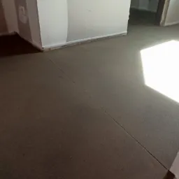 Świeżo wylana, ciemna posadzka w pomieszczeniu z częściowo pomalowanymi ścianami, widoczne szczeliny dylatacyjne i fragment ogrzewania podłogowego w przejściu do kolejnego pomieszczenia.