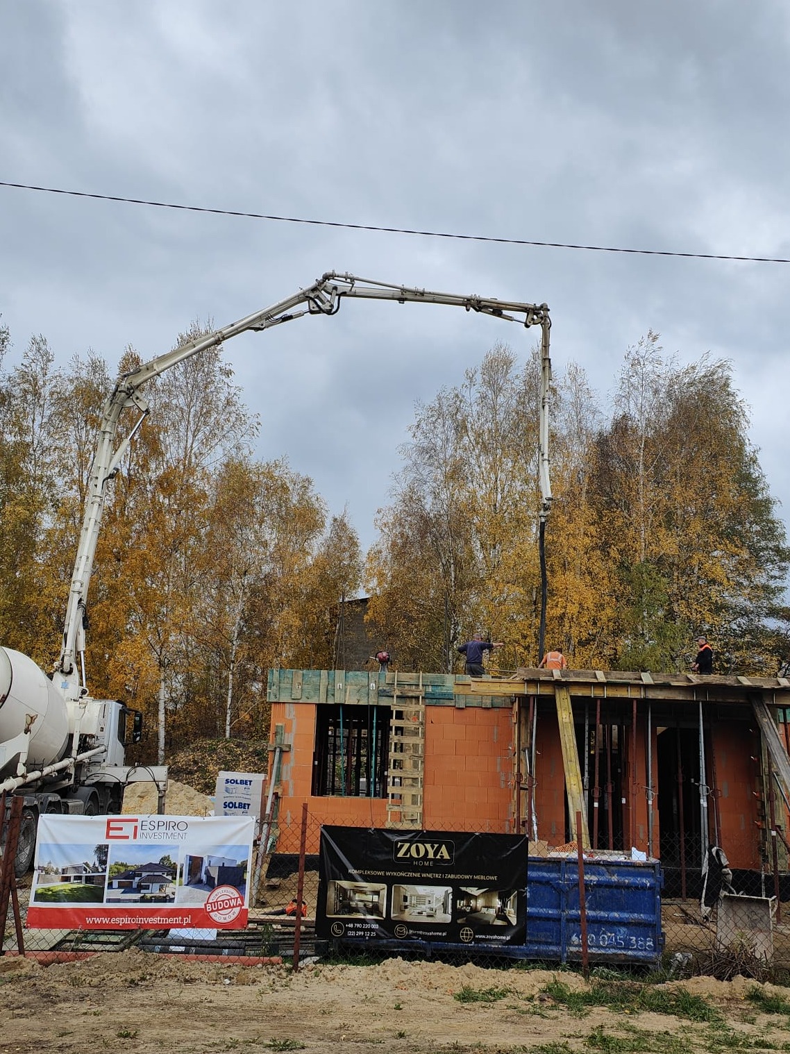 Budowa domu z czerwonej cegły ceramicznej: betonowanie stropu za pomocą pompy do betonu na tle jesiennych drzew, widoczne szalunki i pracownicy.