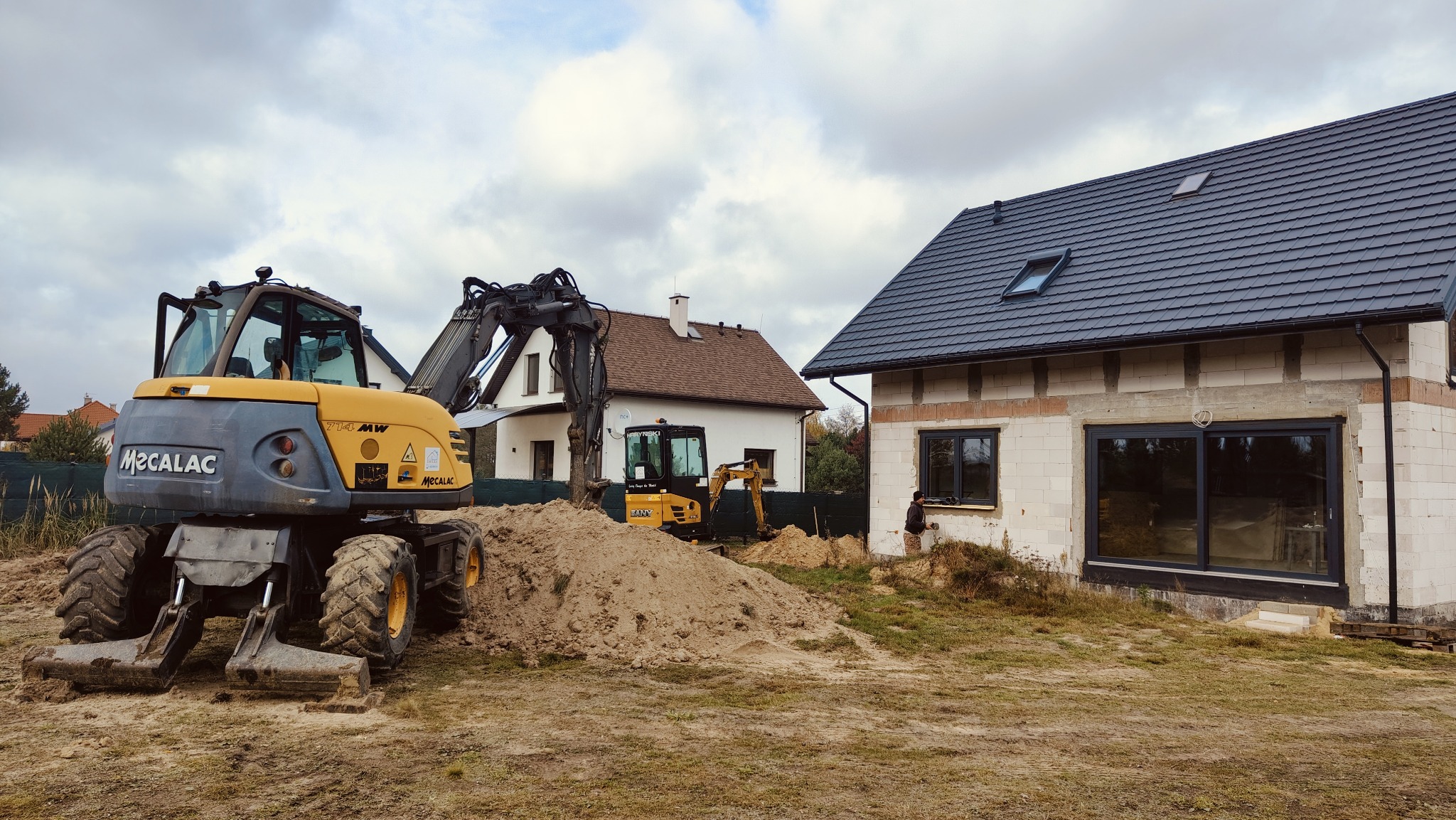 Dwa żółte, kołowe koparki na placu budowy domu z bloczków, w tle drugi dom z brązowym dachem i kominem.