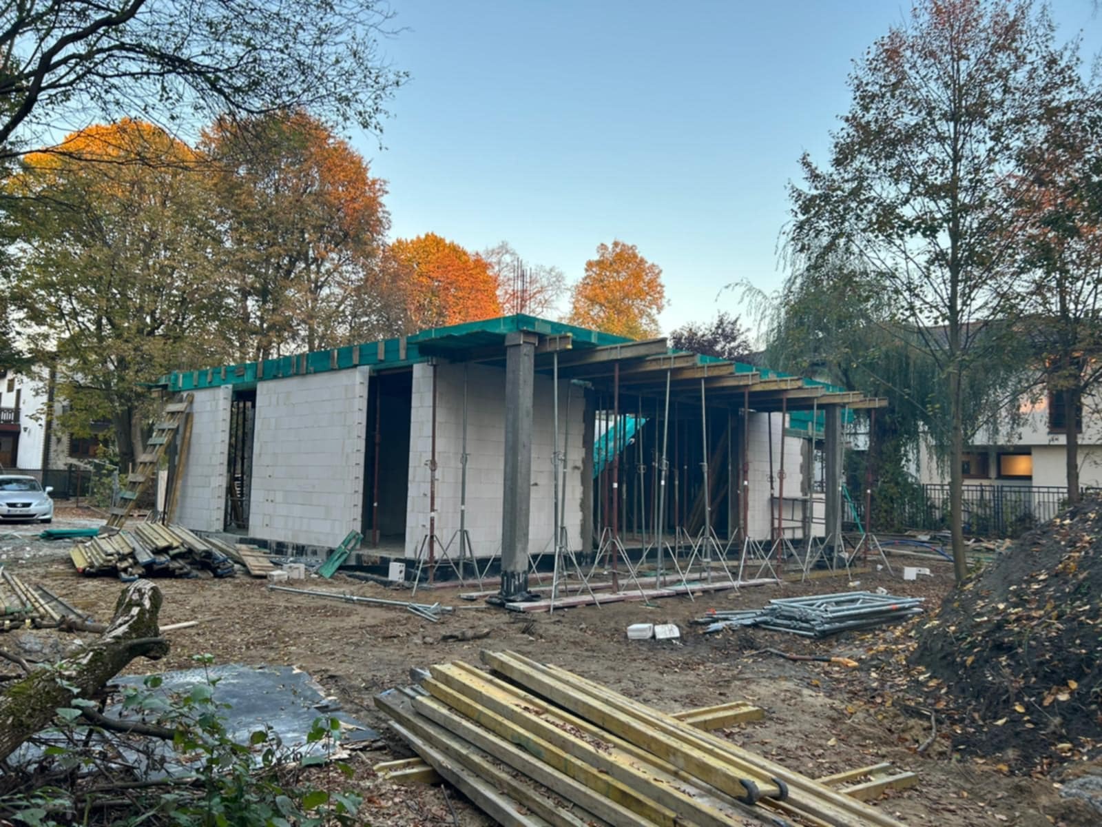 Wznoszenie ścian parterowego domu z bloczków betonowych, widoczne szalunki na stropie, podpory stalowe, teren budowy w jesiennej scenerii z drzewami.