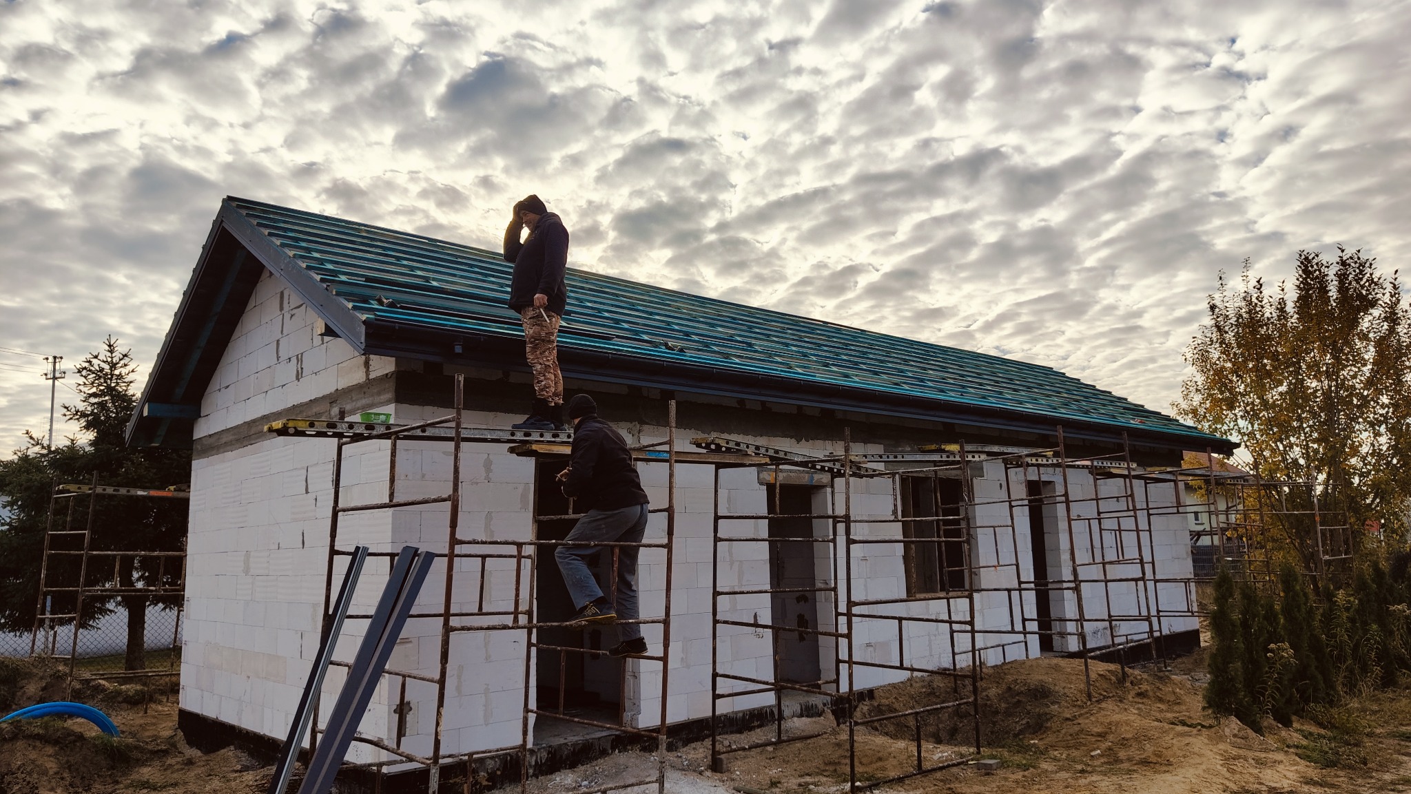 Budowa domu z białych bloczków: dwaj mężczyźni na rusztowaniu montują konstrukcję dachu z zieloną blachą na tle pochmurnego nieba.