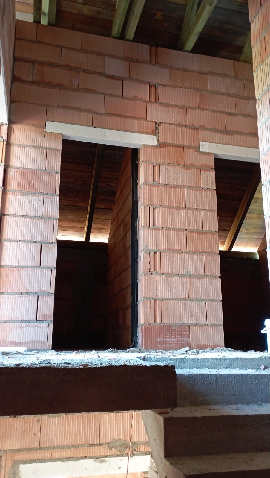 Wnętrze domu w stanie surowym otwartym, widoczne ściany z czerwonej cegły, otwory okienne z nadprożami i drewniana konstrukcja dachu.