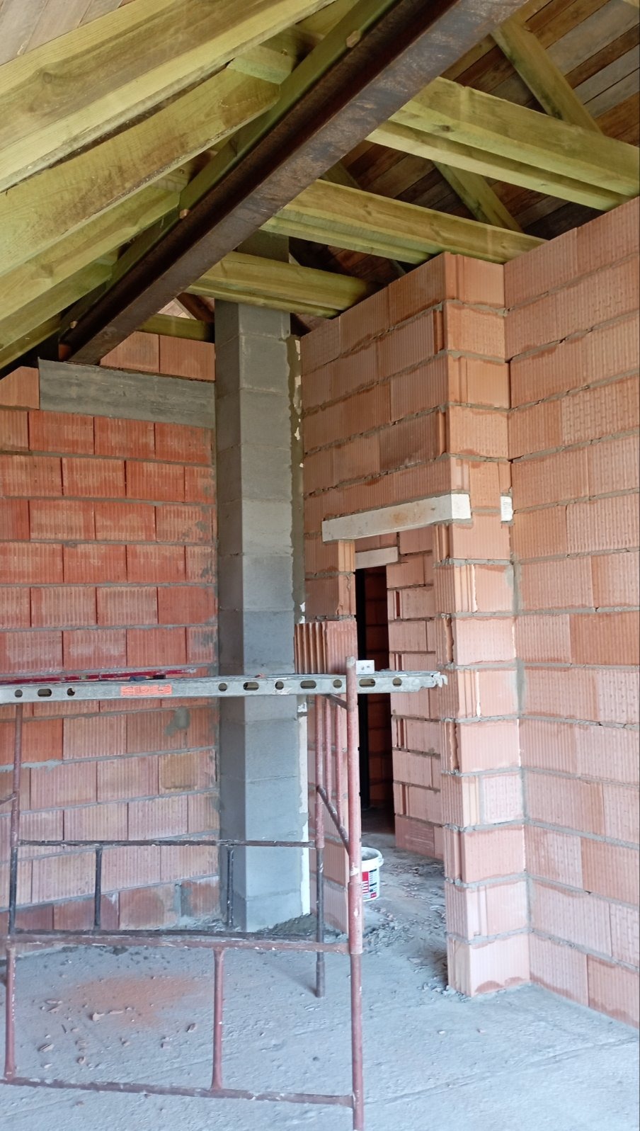 Wnętrze w trakcie budowy: ściany z czerwonej cegły, szara kolumna, drewniany strop, widoczne rusztowanie i pusty otwór drzwiowy.