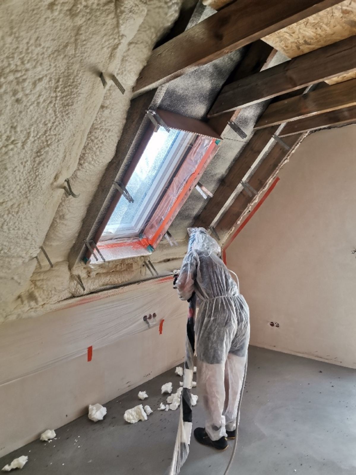 Pracownik w kombinezonie ochronnym natryskuje pianę izolacyjną na poddaszu, widoczne krokwie, okno dachowe zabezpieczone folią, fragment ściany i resztki piany na podłodze.