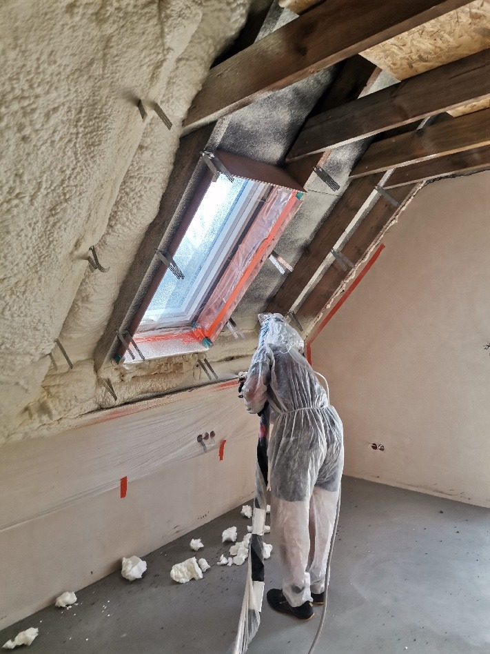 Pracownik w kombinezonie ochronnym aplikuje piankę izolacyjną na poddaszu, widoczne drewniane elementy konstrukcji dachu i okno dachowe.