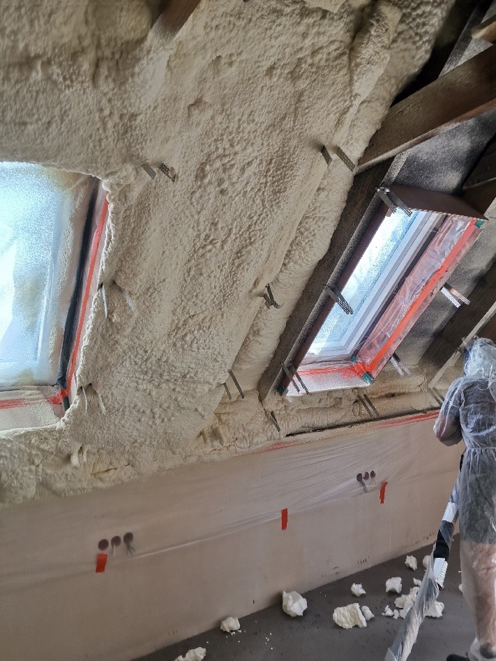 Poddasze ocieplone pianką, widoczne okna dachowe i folia ochronna na ścianie, fragment drabiny i osoba w kombinezonie ochronnym.