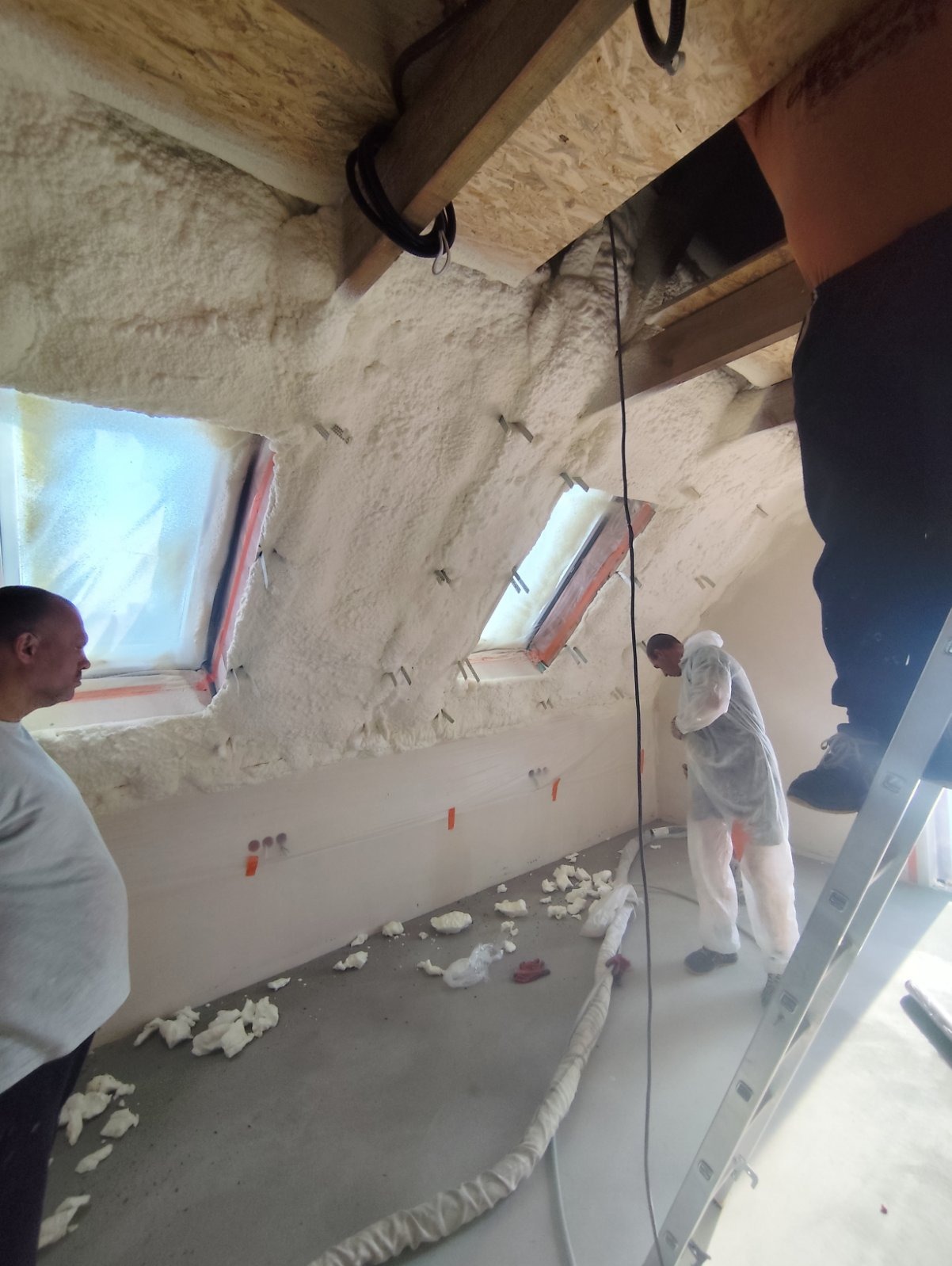 Poddasze w trakcie ocieplania: aplikacja białej piany na skośne ściany z widocznymi oknami dachowymi, pracownicy w kombinezonach ochronnych, fragmenty piany na podłodze, drabina i elementy...