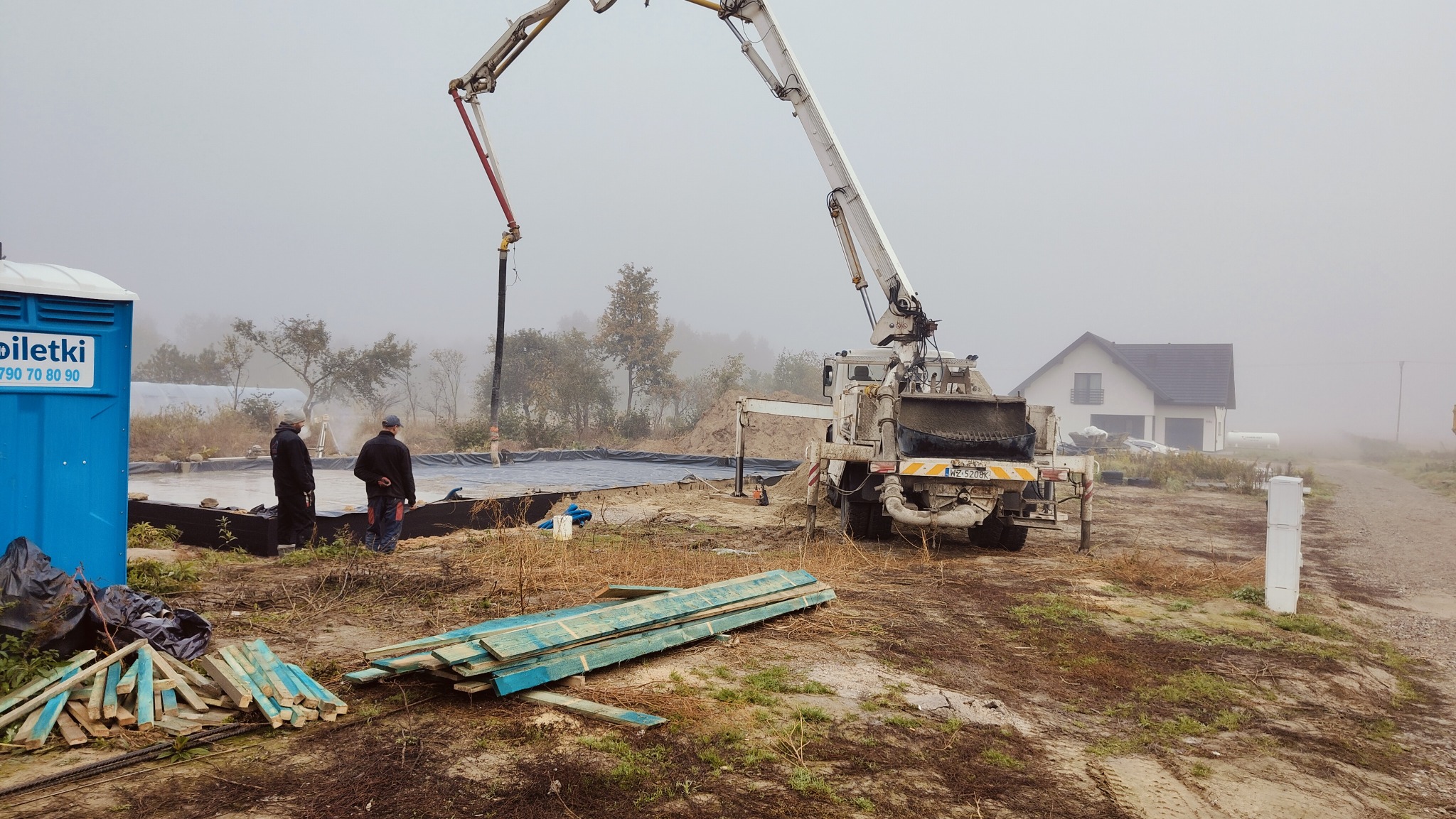 Wylewanie betonu do przygotowanego wykopu pod fundamenty domu jednorodzinnego, widoczny dźwig z pompą do betonu, dwaj pracownicy kontrolują proces, w tle budynek w budowie i przenośna toaleta.