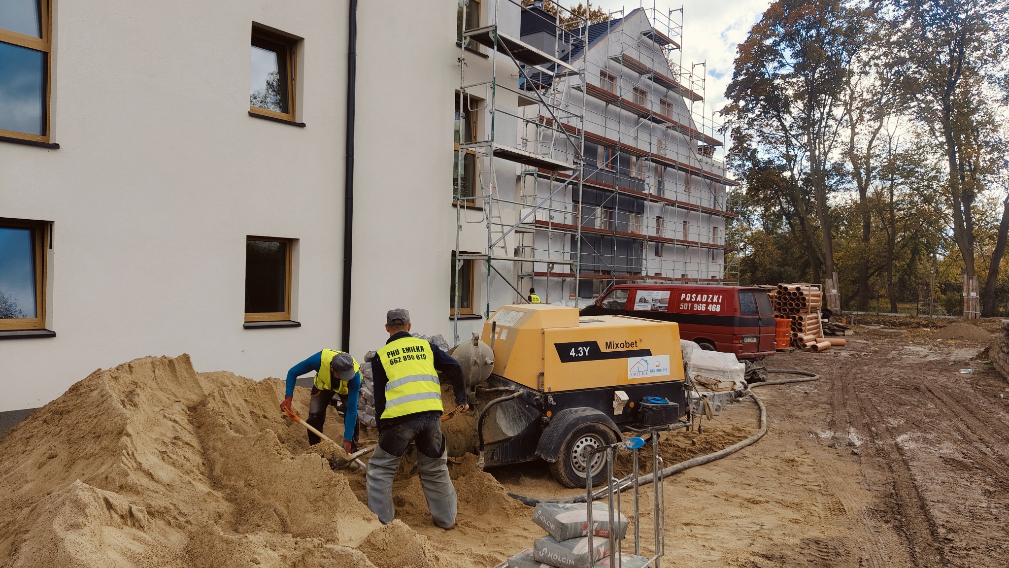 Pracownicy budowlani łopatami przerzucają piasek obok pompy do wylewek Mixobet 4.3Y na placu budowy, w tle budynek w trakcie ocieplania ze stojącymi rusztowaniami i zaparkowany bus z reklamą posadzek.