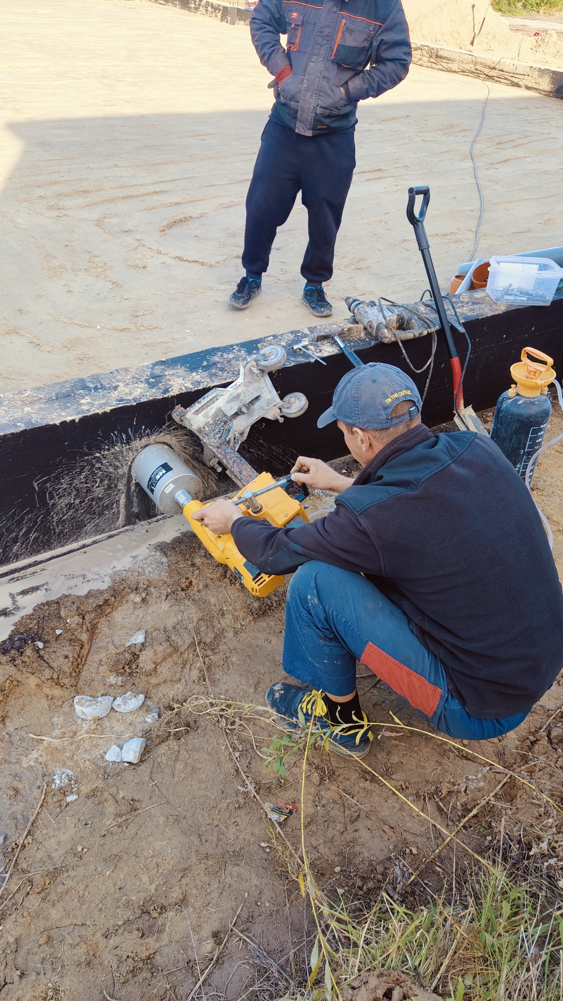 Pracownik w trakcie wykonywania otworu w elemencie konstrukcyjnym przy użyciu wiertnicy diamentowej, widoczny fragment fundamentu i drugi pracownik w tle.