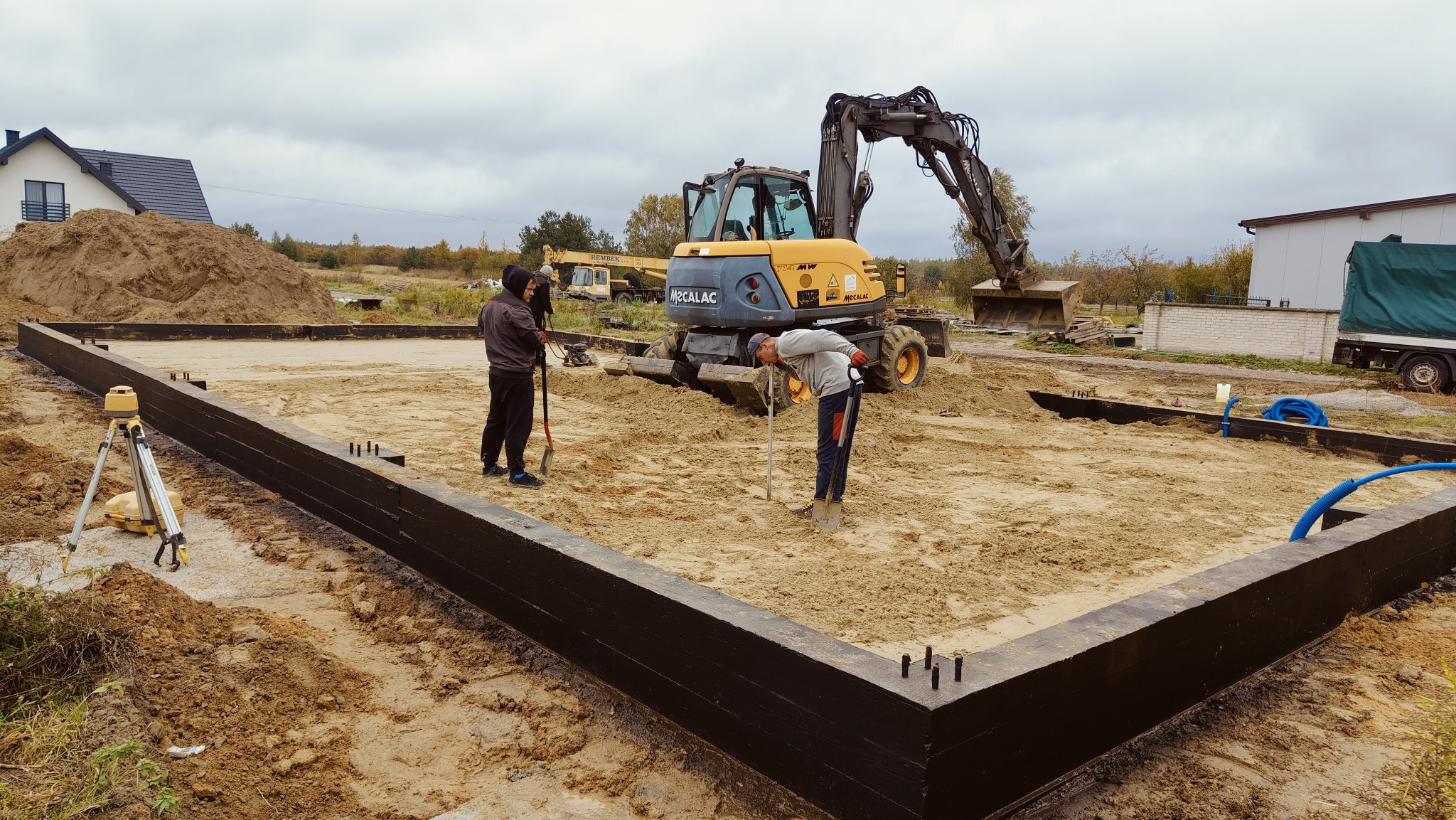 Przygotowanie terenu pod fundamenty domu jednorodzinnego z użyciem mini koparki Mecalac i niwelatora laserowego, widoczne drewniane szalunki pomalowane na czarno i wystające pręty zbrojeniowe.