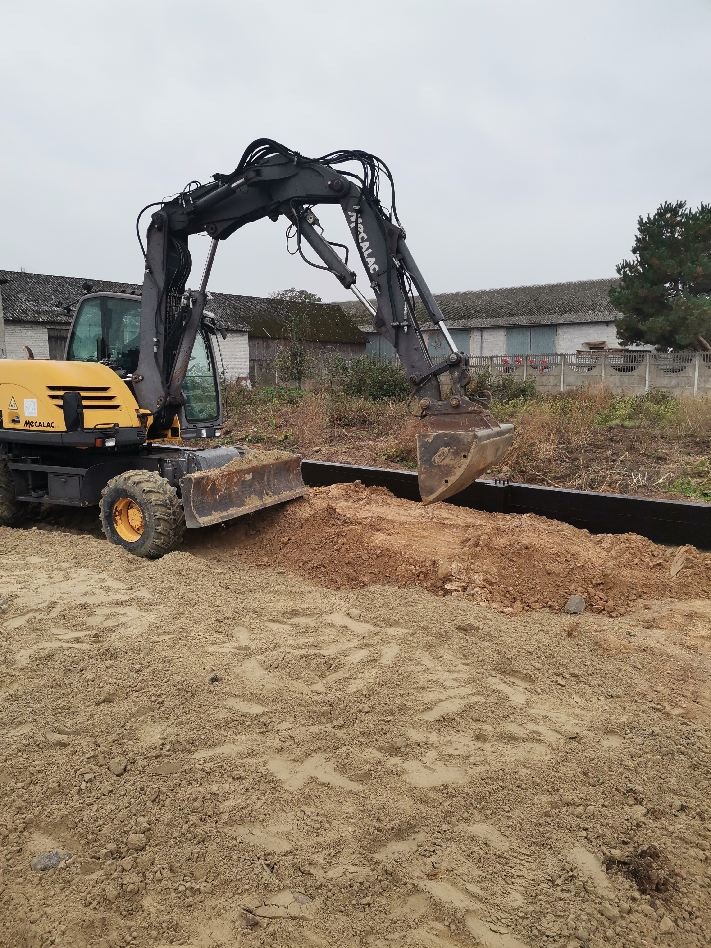 Żółto-czarna koparka Mecalac wyrównuje teren pod budowę, widoczne przygotowane czarne szalunki, w tle zabudowania gospodarcze.