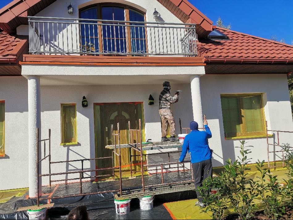 Dwaj pracownicy na rusztowaniu nanoszą tynk na elewację domu jednorodzinnego z zabezpieczonymi oknami i drzwiami, widoczna kolumna i balkon z metalową balustradą.