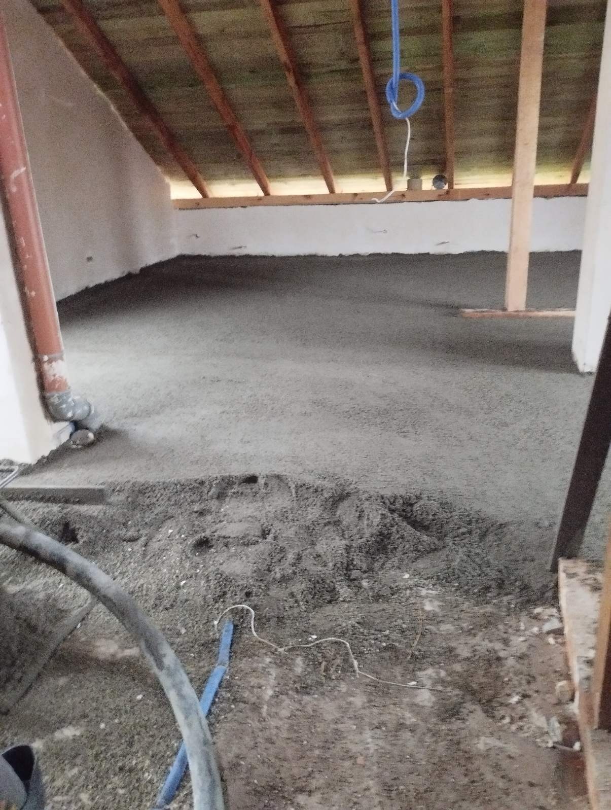Świeżo wylana, mokra, szara masa na podłodze poddasza, widoczny wąż do pompowania mieszanki i drewniany szkielet dachu.