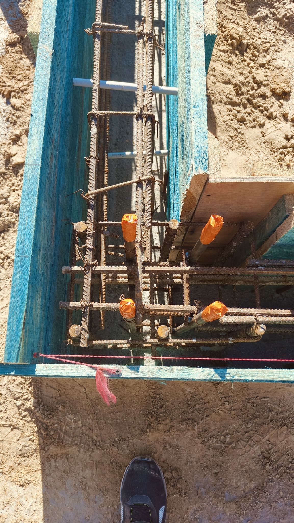 Zbrojenie fundamentu w szalunku z niebieskiego drewna, zabezpieczone rury w pomarańczowych osłonach, widoczny fragment buta pracownika.