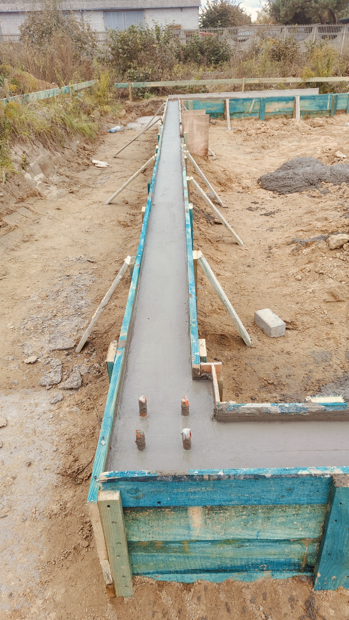 Wylany beton w szalunku fundamentowym z wystającymi prętami zbrojeniowymi, widoczne podpory i przygotowany grunt wokół.