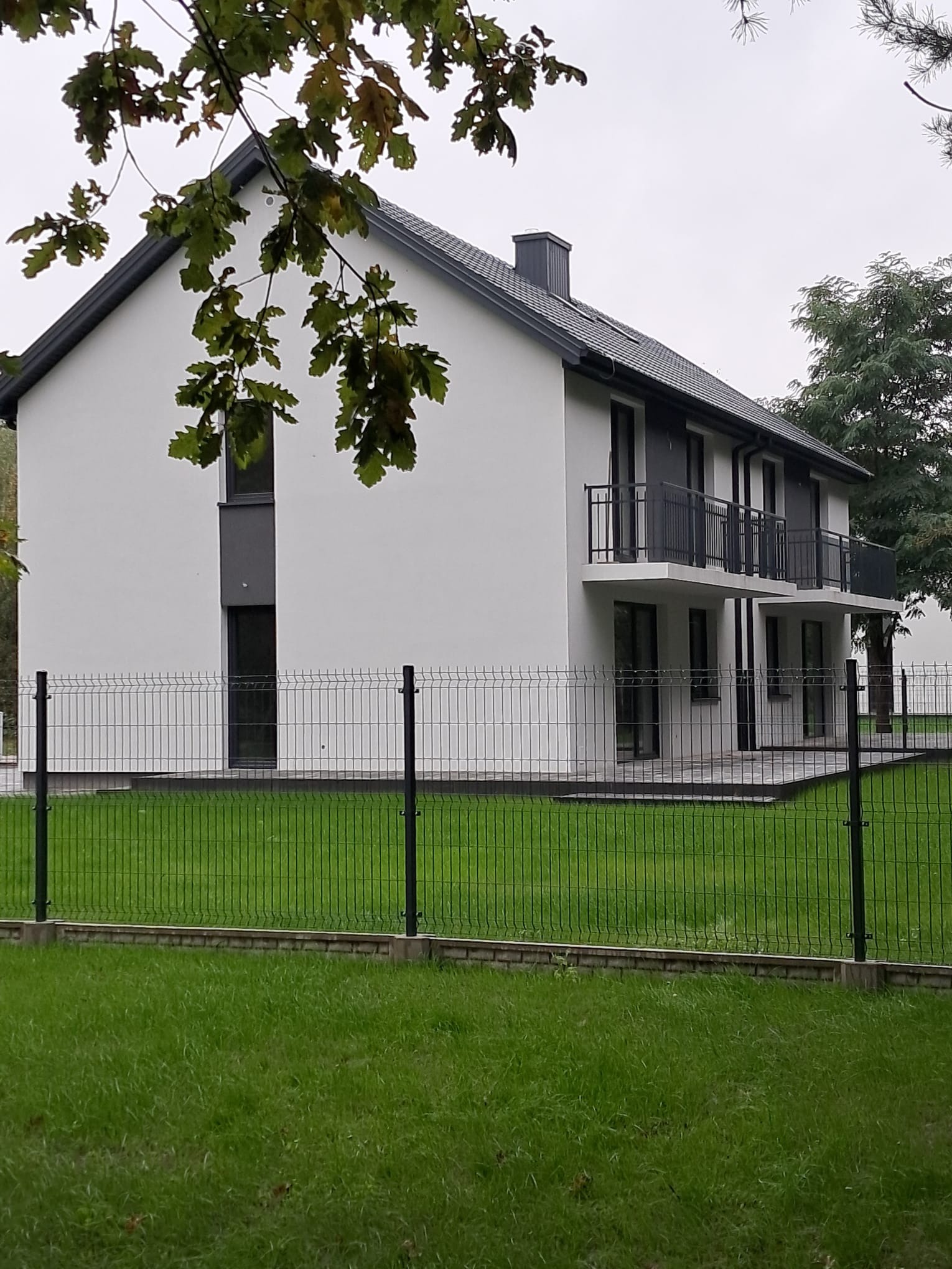 Dwukondygnacyjny dom z białymi ścianami i ciemną stolarką okienną oraz balkonami, ogrodzony metalową siatką na podmurówce, widok z zewnątrz w pochmurny dzień.