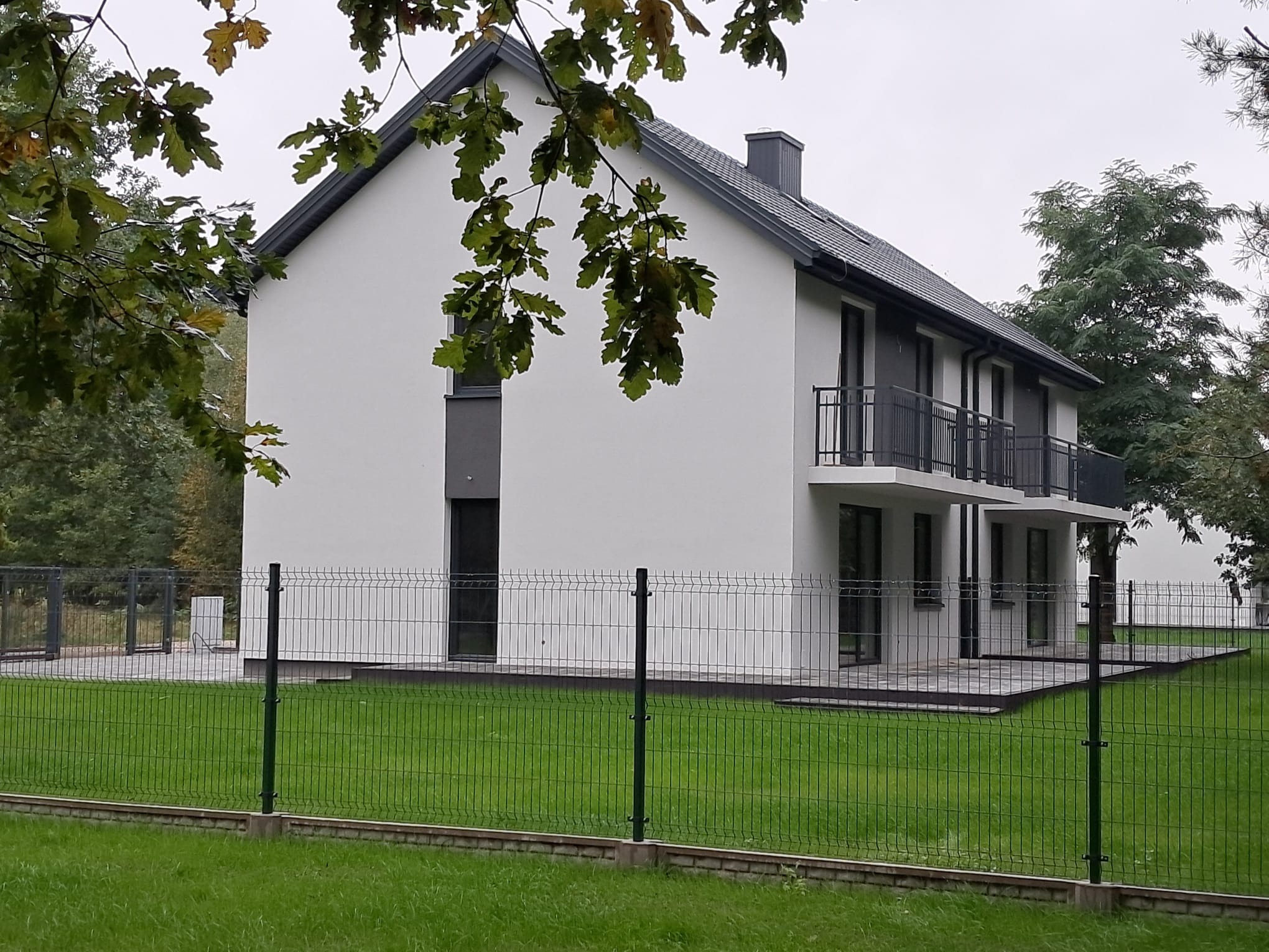 Nowoczesny dom jednorodzinny z białymi ścianami i szarymi elementami wykończeniowymi, widoczny zza metalowego ogrodzenia i zielonego trawnika, z fragmentami drzew w tle.