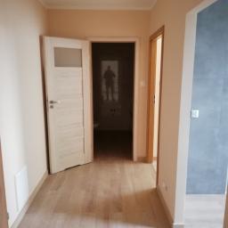 MAR-BUD Uslugi Budowlane Mariusz Szymulewski - Korytarz z drewnopodobną podłogą, świeżo pomalowanymi ścianami w odcieniach beżu i otwartymi drzwiami do łazienki z widocznym odbiciem osoby w lustrze.