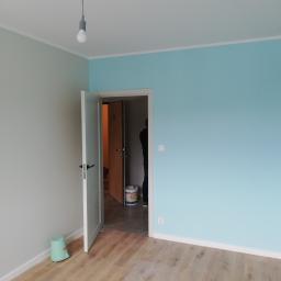 MAR-BUD Uslugi Budowlane Mariusz Szymulewski - Pokój po malowaniu ścian na dwa kolory: błękitny i szary, z widoczną otwartą białą futryną drzwi, drewnopodobną podłogą i wiadrem po farbie w głębi pomieszczenia.