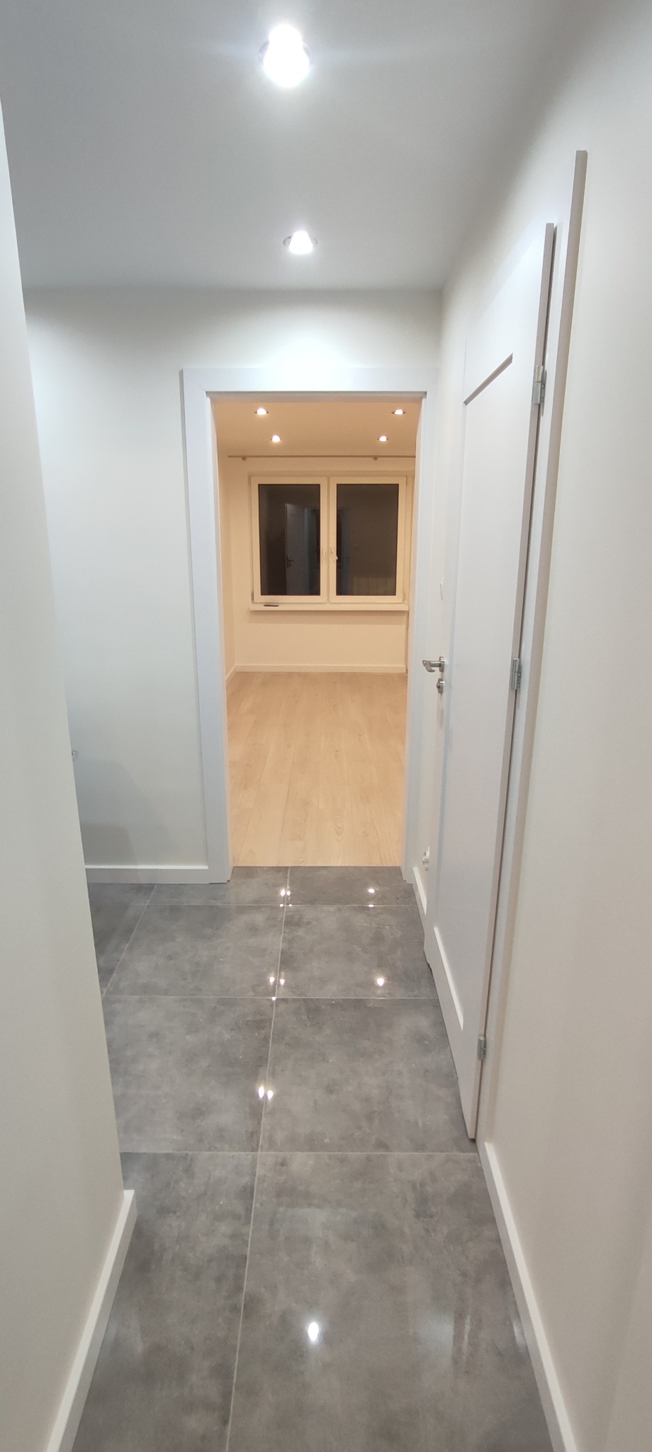 Korytarz z szarą, połyskującą podłogą z płytek, białe ściany i drzwi, w tle widoczny pokój z drewnianą podłogą i oknem.