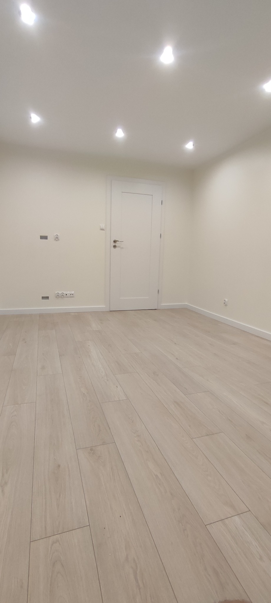 Pusta, jasna przestrzeń z panelami podłogowymi imitującymi drewno, białe ściany z gniazdkami i włącznikami, białe drzwi wewnętrzne oraz oświetlenie punktowe na suficie.