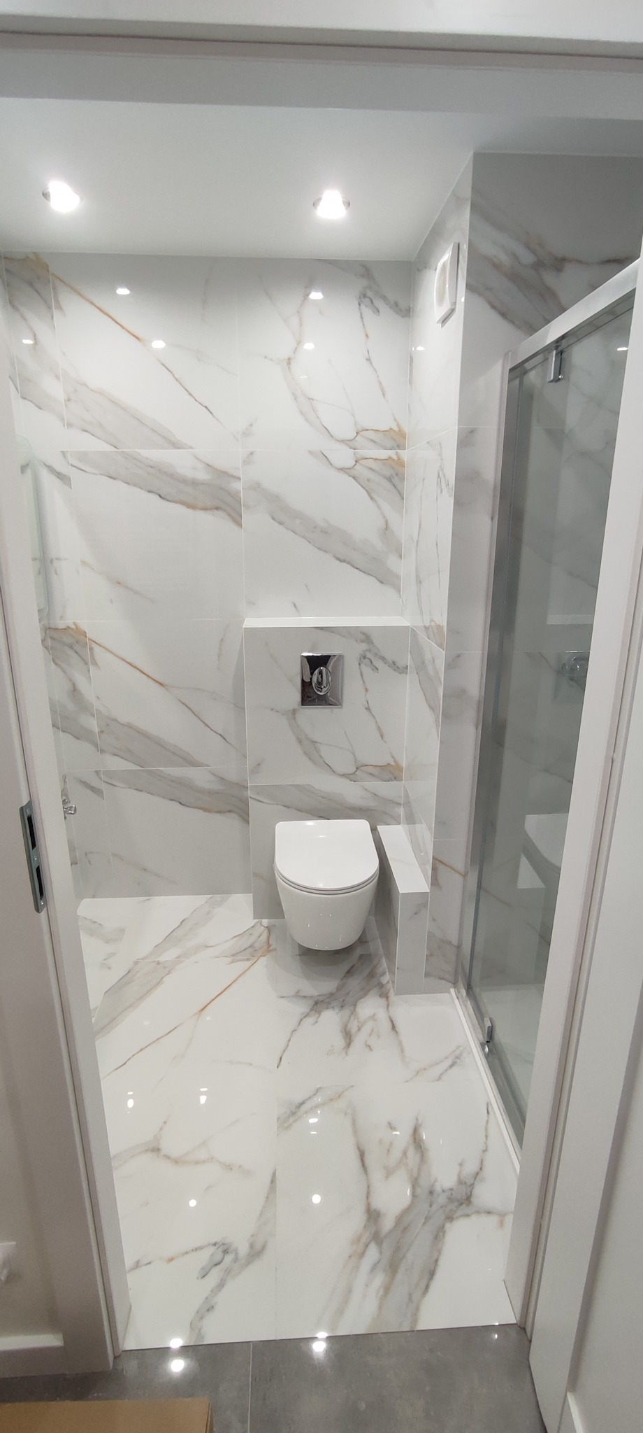 Nowoczesna łazienka z białymi płytkami imitującymi marmur, podwieszana toaleta z chromowanym przyciskiem i szklana kabina prysznicowa.