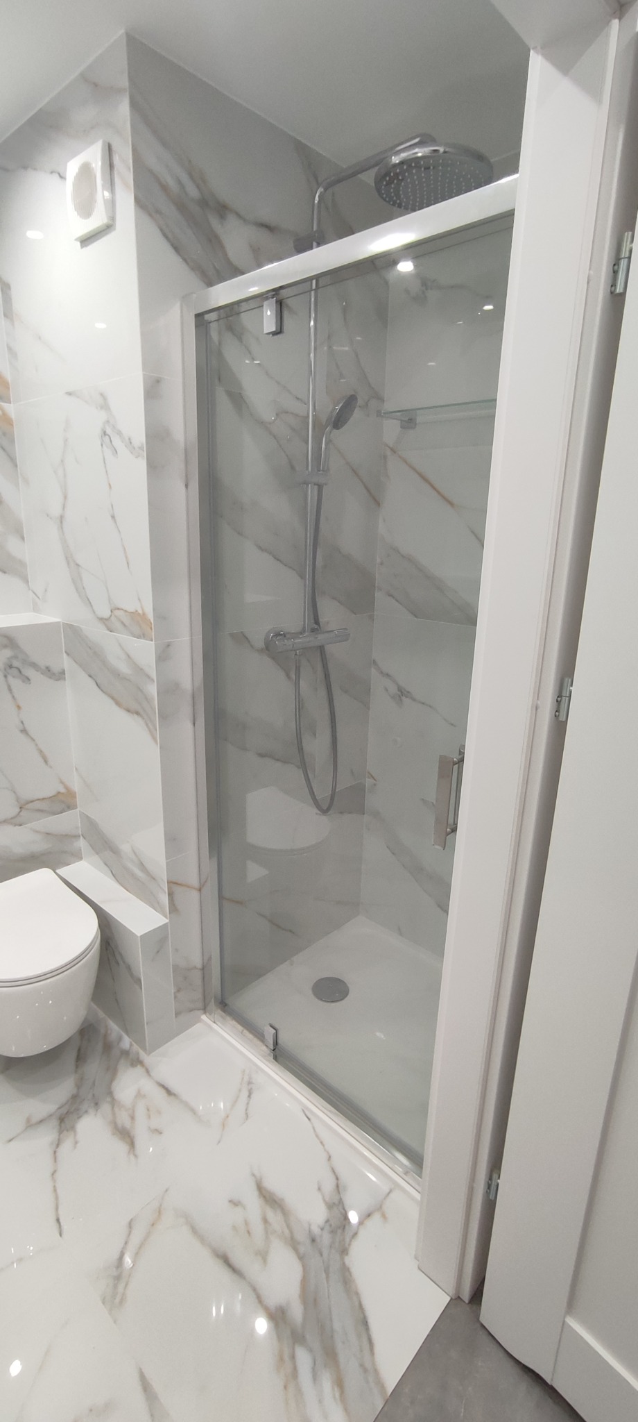 Nowoczesna kabina prysznicowa z deszczownicą i baterią termostatyczną, obok toaleta i płytki imitujące marmur na ścianach i podłodze.