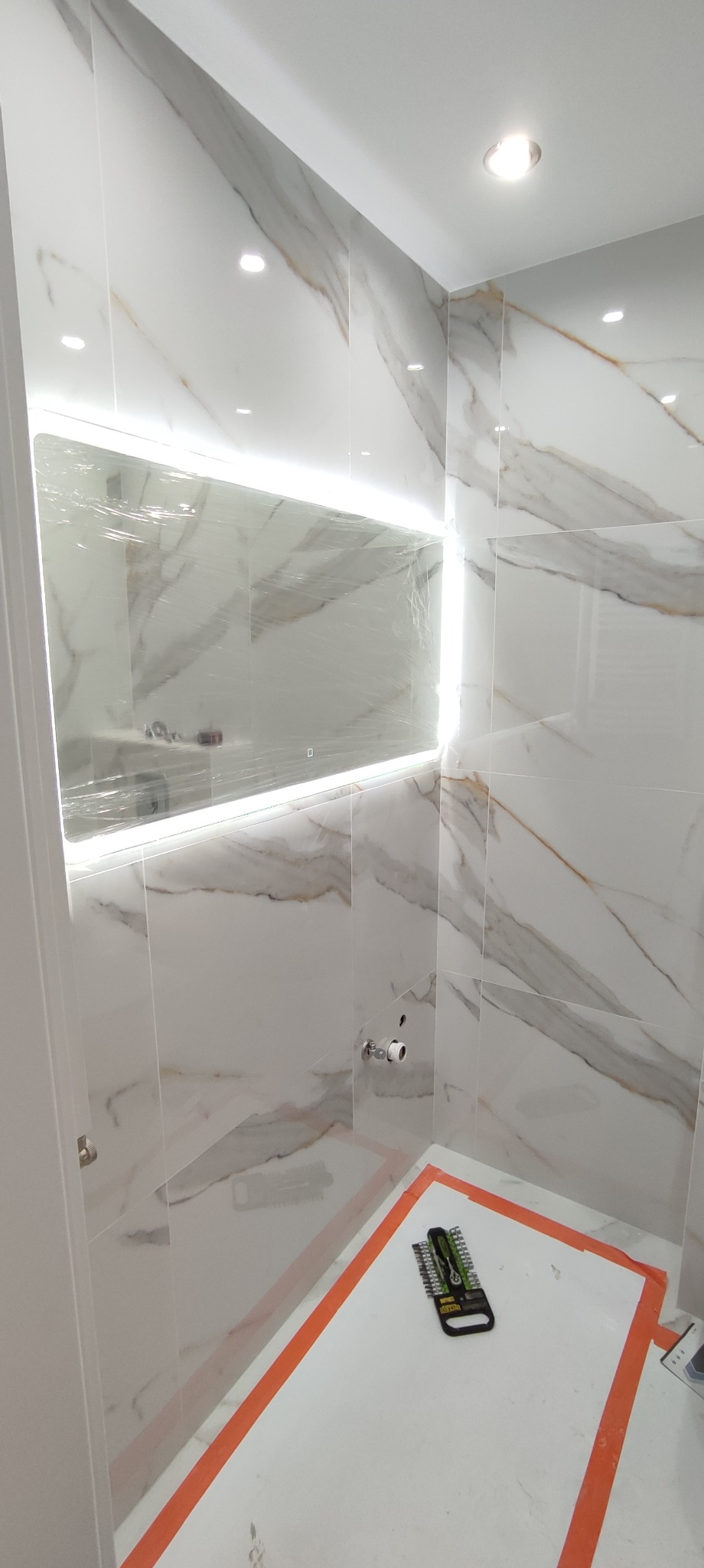 Łazienka w trakcie remontu, białe płytki imitujące marmur na ścianach, podświetlane lustro zabezpieczone folią, narzędzia glazurnicze na podłodze zabezpieczonej folią malarską.