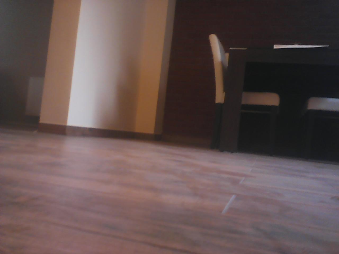 Ukończona podłoga w odcieniach brązu, z widocznymi panelami i ciemnym stołem z krzesłami w tle. Perspektywa z niskiego kąta, blisko podłogi.