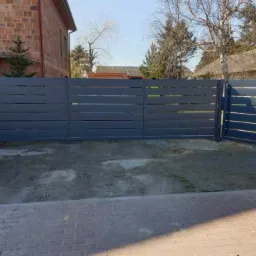 Nowoczesne, poziome przęsła grafitowego ogrodzenia z metalu, widoczne z zewnątrz posesji, z ceglanym budynkiem w tle i fragmentem kostki brukowej na pierwszym planie.