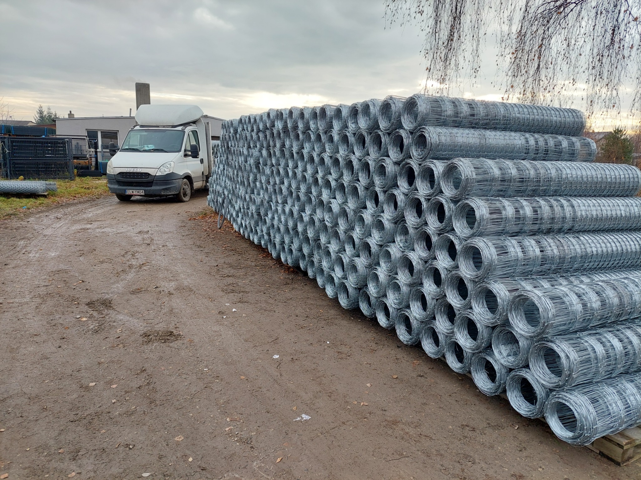 Stos zwojów siatki ogrodzeniowej ułożony na ziemi obok białego samochodu dostawczego na tle pochmurnego nieba i drzew.