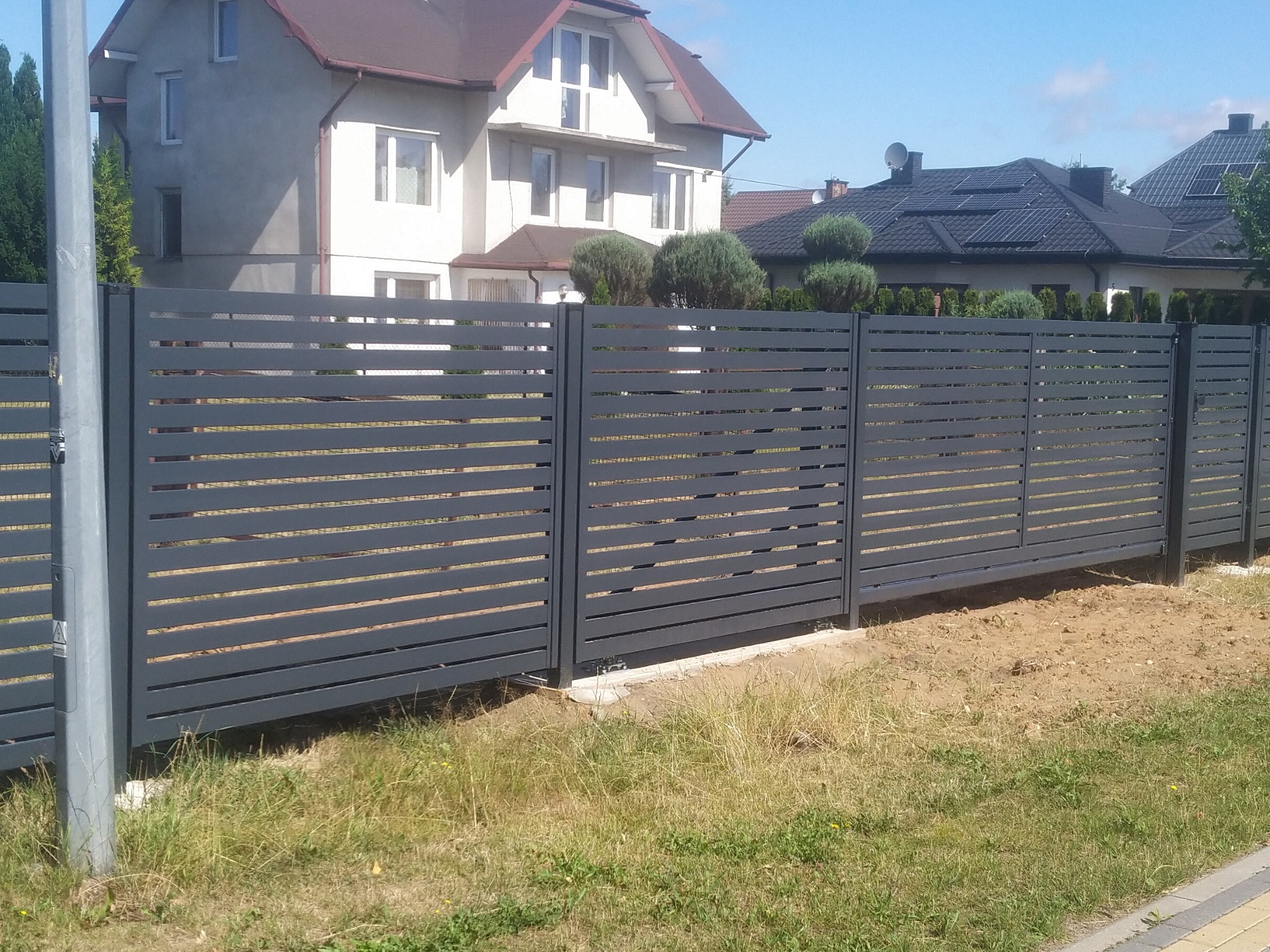 Nowoczesne, poziome, szare przęsła ogrodzenia metalowego na tle dwóch domów jednorodzinnych w słoneczny dzień.