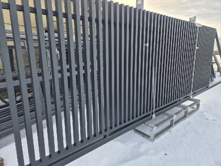 Nowoczesne, szare ogrodzenie posesji z pionowych, metalowych profili. Widoczne elementy konstrukcyjne i zimowy krajobraz w tle. Minimalistyczny design.