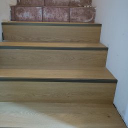 Mirosław Brzozowski - Wykończone schody z panelami o odcieniu jasnego dębu i szarymi listwami. Widoczne niedokończone płytki na górnych stopniach, co sugeruje trwający remont.
