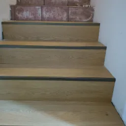 Wykończone schody z panelami o odcieniu jasnego dębu i szarymi listwami. Widoczne niedokończone płytki na górnych stopniach, co sugeruje trwający remont.