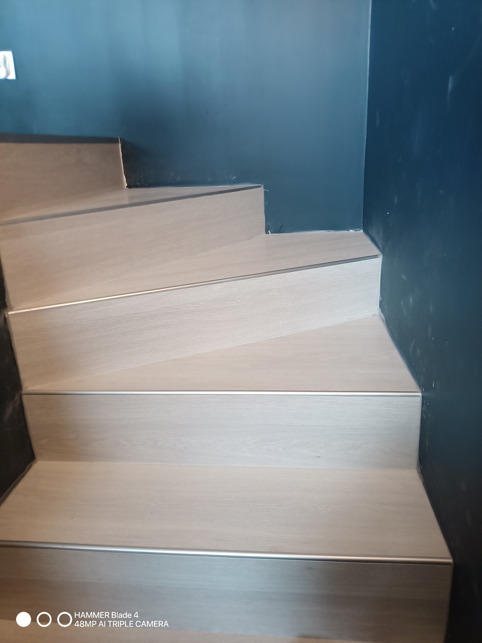 Schody z panelami w odcieniach beżu, wykończone aluminiowymi listwami, na tle ściany w kolorze ciemnego granatu. Widok z dołu.