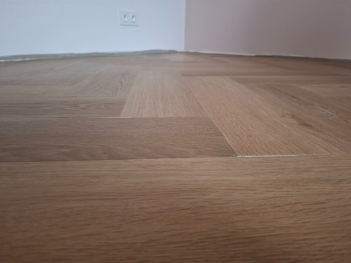 Ułożony parkiet w jodełkę francuską, widoczne gniazdko elektryczne na ścianie. Jasne drewno, delikatne oświetlenie.