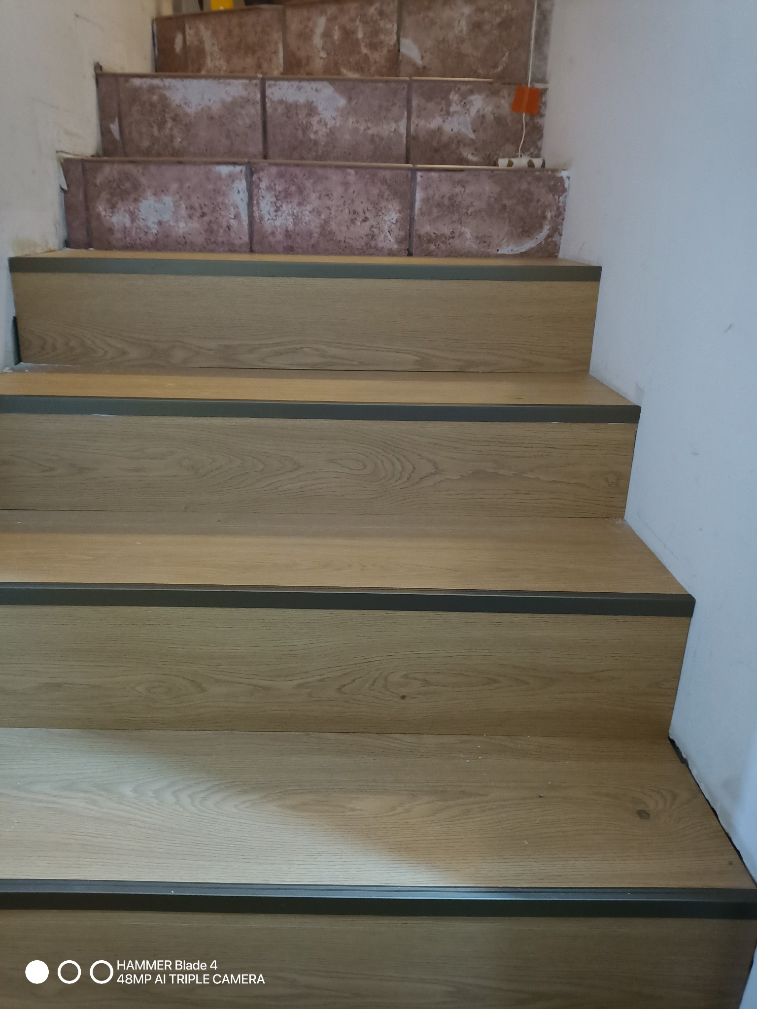 Wykończone schody z panelami o odcieniu jasnego dębu i szarymi listwami. Widoczne niedokończone płytki na górnych stopniach, co sugeruje trwający remont.